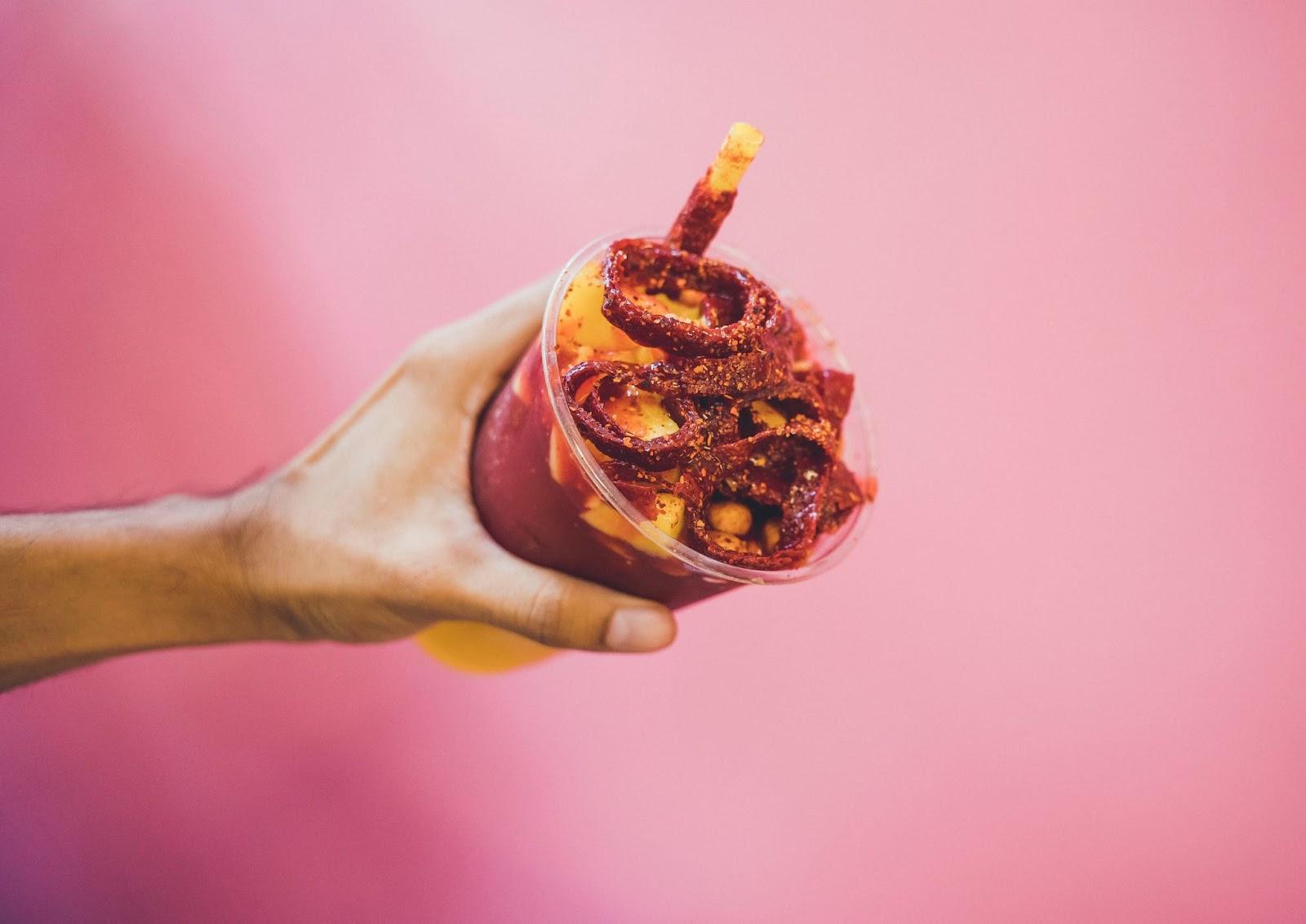 A dismembered hand holds a plastic cup of mango slushy with a pink wall in the background.