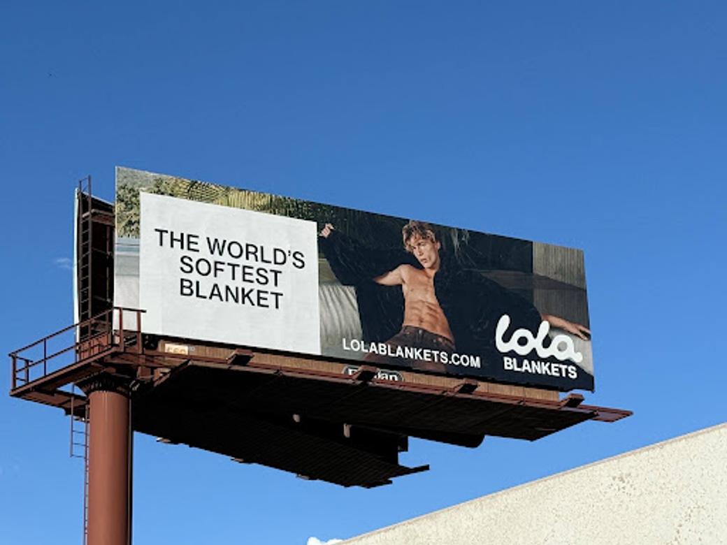 A billboard of Lola Blankets showing a white man shirtless, abs exposed but wrapped in a black blanket marketing “the world’s softest blanket.”