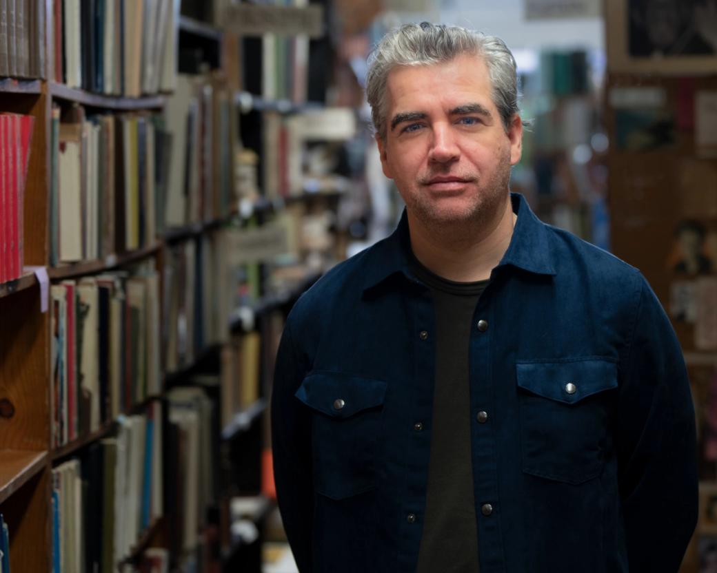 A man in a blue shirt standing next a bookshelf.