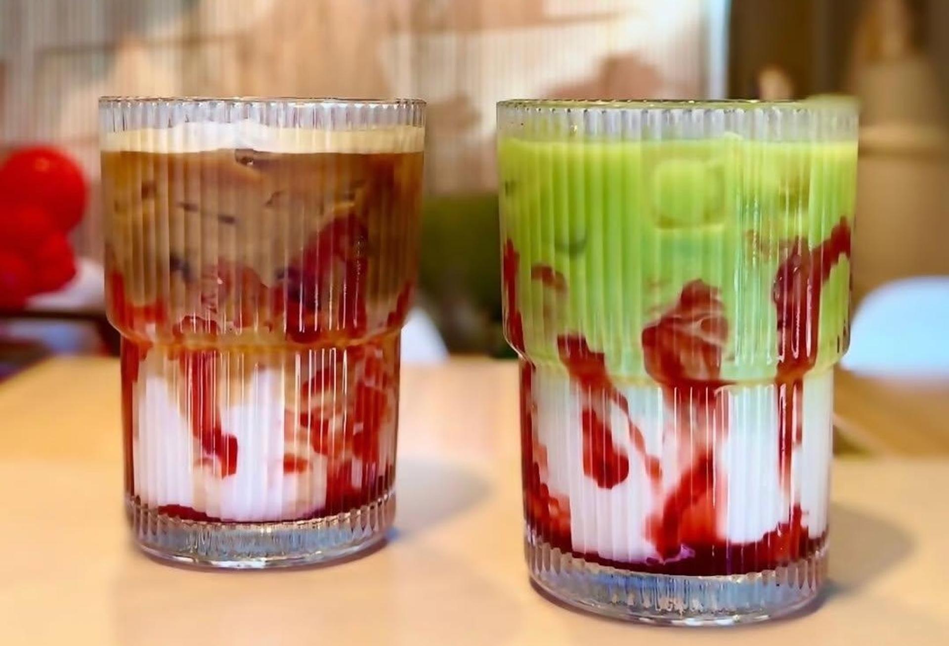 On a tabletop sit two pleated glasses next to one another, both filled with iced coffee drinks. One is topped with brown espresso and the other with green matcha, but both share streaks of red raspberry meant to resemble blood, as well as a white mocha base. 