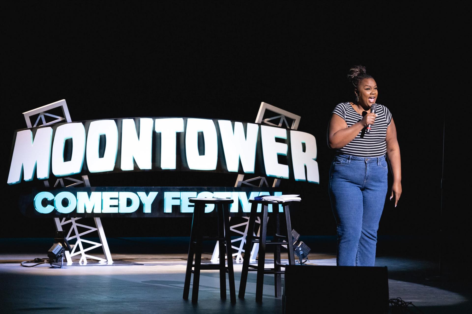 Nicole Byer speaking into a microphone in front of a sign that says "Moontower Comedy Festival."