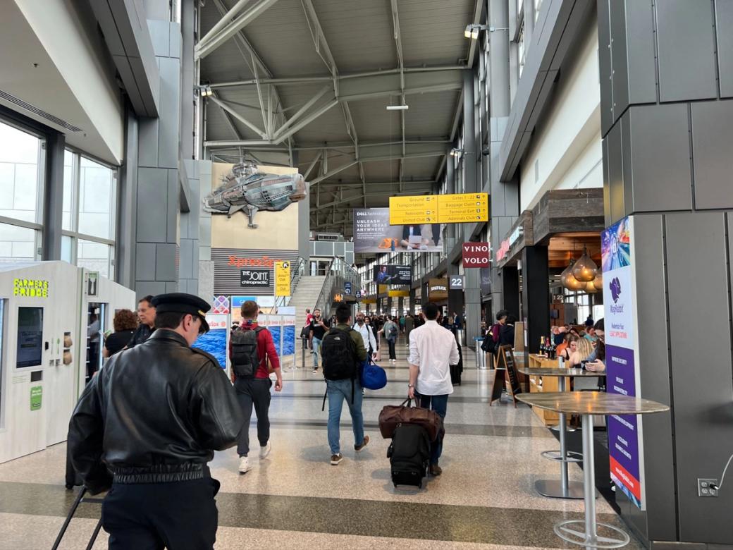 People carrying luggage walk down a busy corridor at AIBA. Signs point to locations of different amenities.