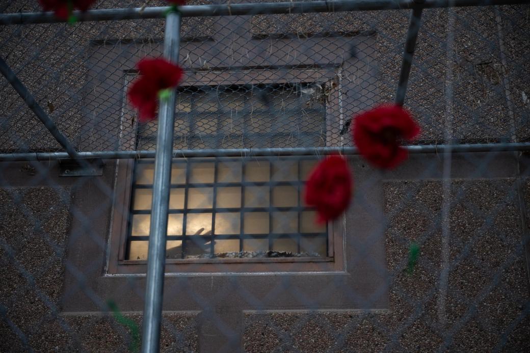 Red poppies are hung on the fence in the foreground as a detainee places their hand against a window of the Aurora ICE Processing Center in the background.