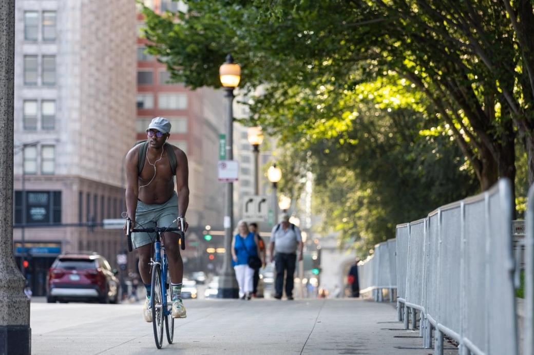 A shirtless man on a bike riding on a sidewalk. It's a sunny day. The sun is covered by trees