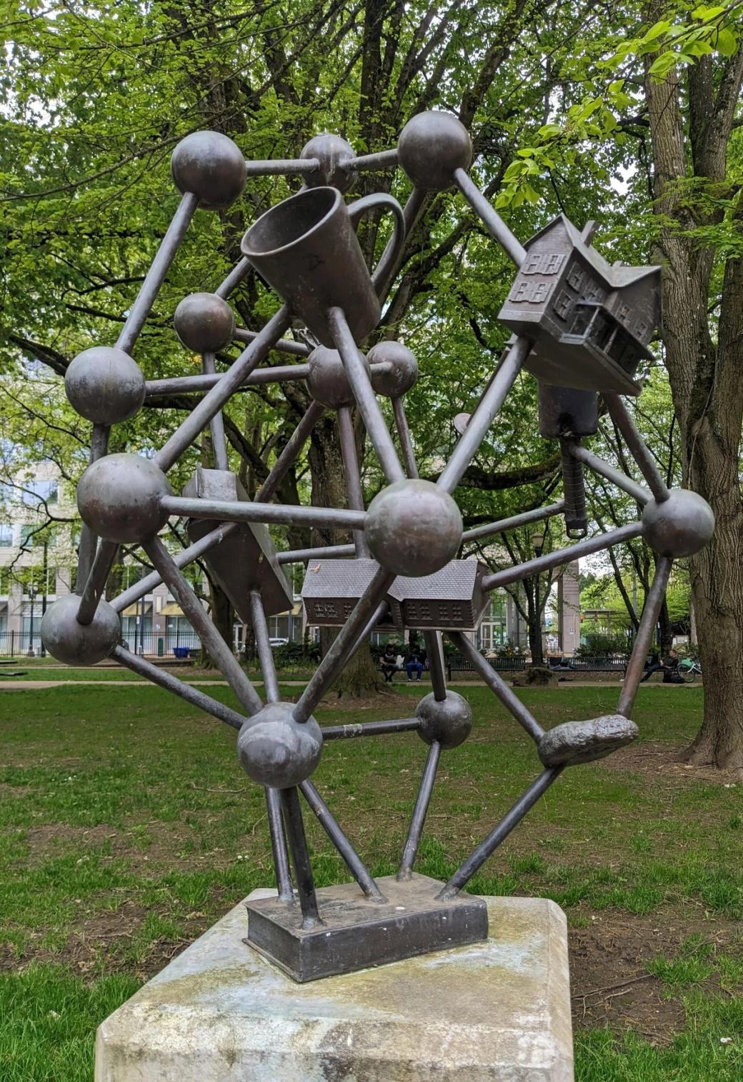 Metal sculpture that looks like a model of a molecule but in place of some atoms are a house, a coffee cup, a milk carton, and school.