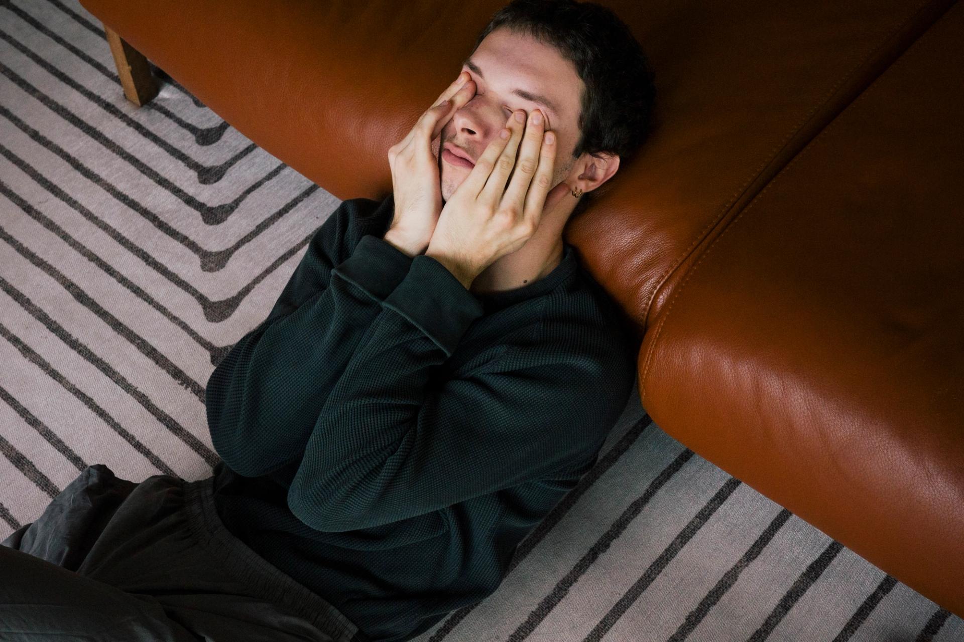 A man sits on a couch rubbing his eyes with his hands.