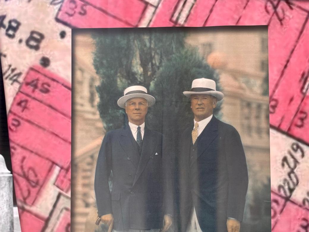 a photo of two middle-aged white men in dark blue suits and white hats with a background photo of a closeup pink city planning map
