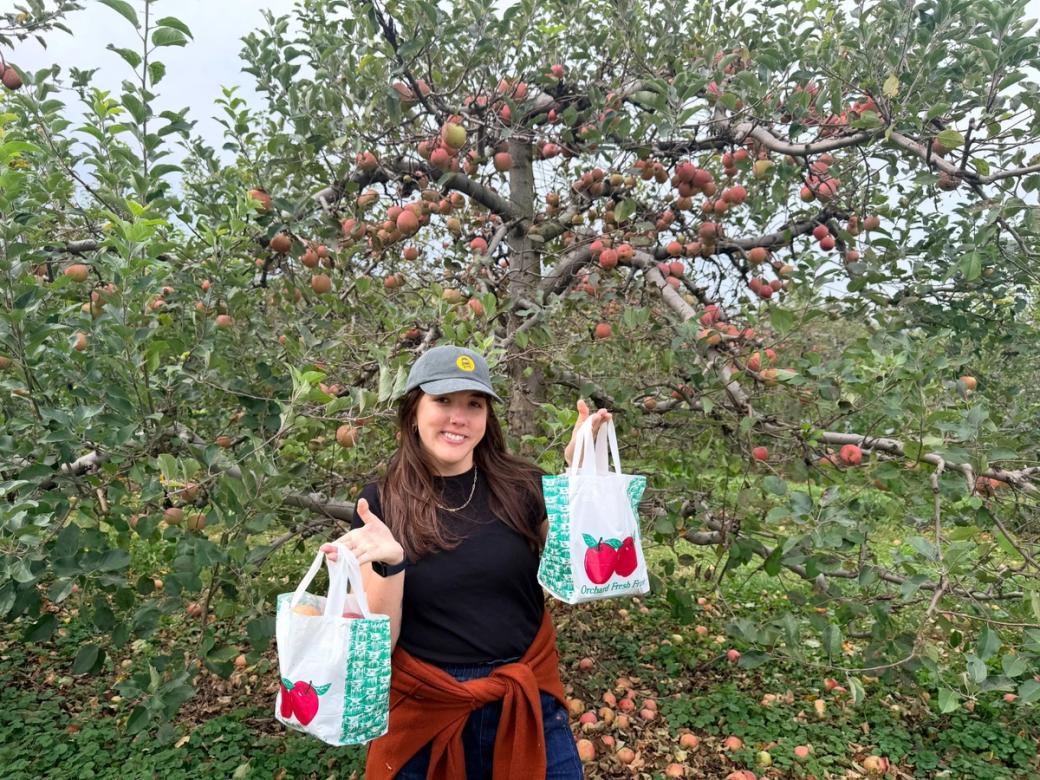 A smiling woman in a gray baseball cap and black shirt holds up two bags of freshly picked apples in an orchard, surrounded by trees filled with red and green apples.
