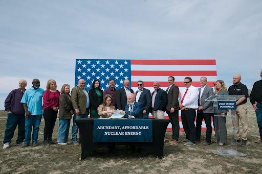 Utah Gov. Spencer Cox signs a memorandum in Tooele County, surrounded by state officials and industry representatives, with a large American flag backdrop.