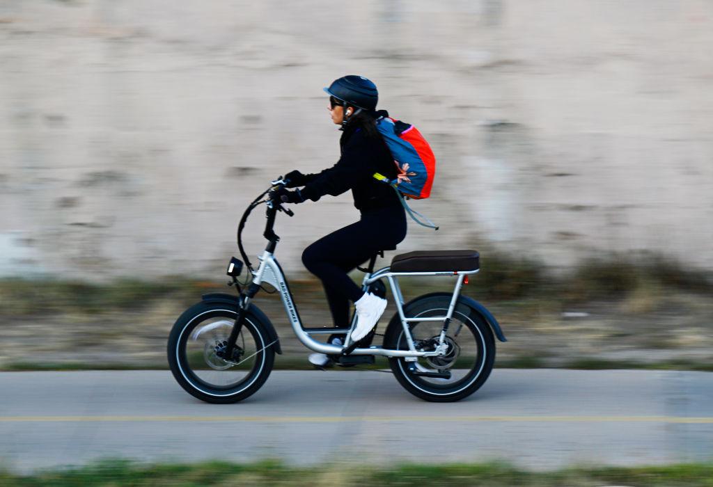 A person wearing a helmet, black clothing, and a colorful backpack rides an e-bike along a path, captured with motion blur conveying speed.