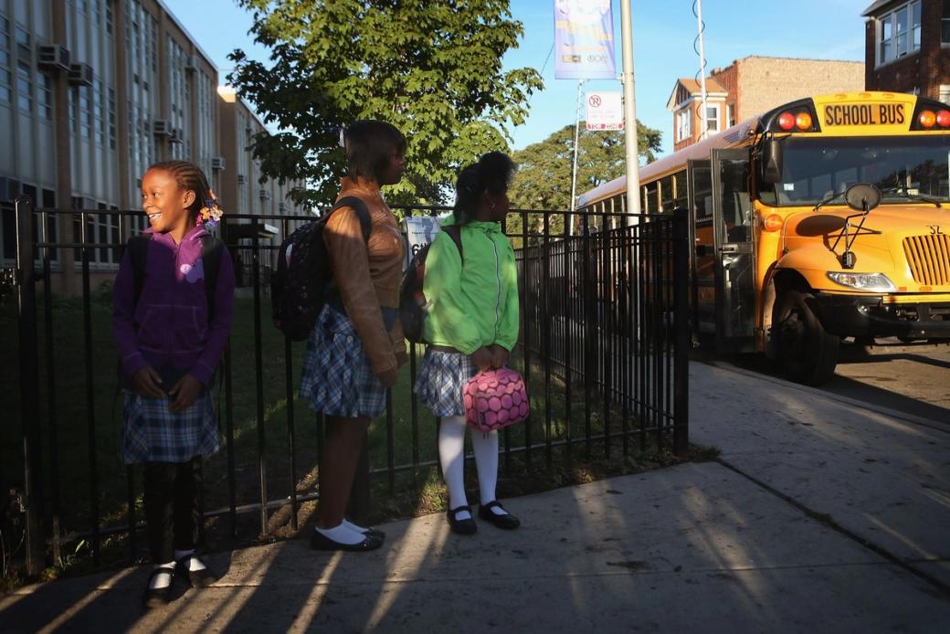 Three girls waiting in line by a gate for a school bus. The school bus is pulling up now