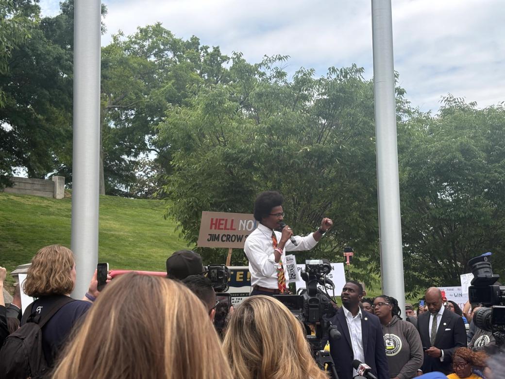 State Rep. Justin Pearson, a Black man in a white button-up and red tie, speaks outside into a microphone in front of protestors and media with cameras. 
