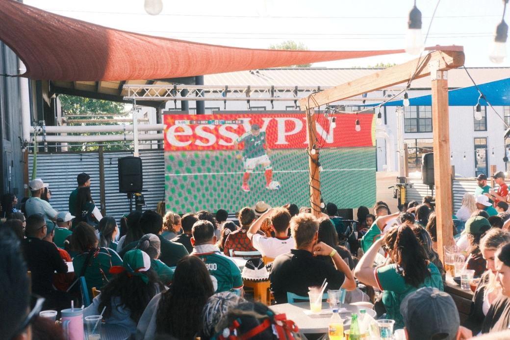 crowd in Mexico jerseys on an outdoor patio watching a large screen