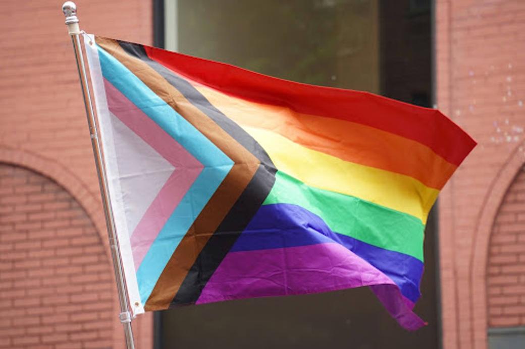 The Progress Pride flag representing the LGBT community. There are horizontal red, orange, yellow, green, blue, and purple stripes with triangular stripes in black, brown, light blue, pink, and white pointing right from the flag pole