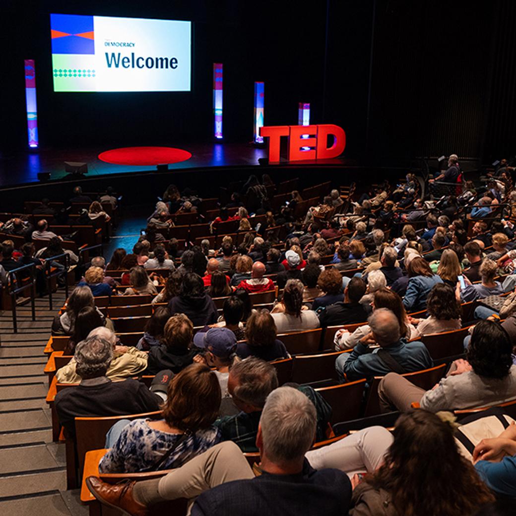 Audience seated in a dimly lit theater facing a stage with a TED logo and "Welcome" on a screen, creating an anticipatory atmosphere.