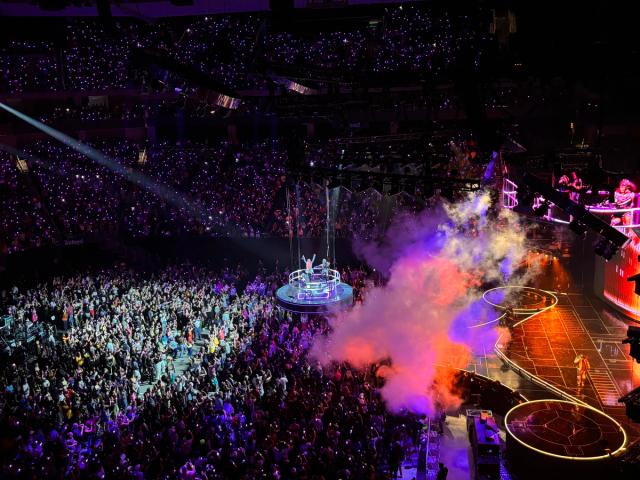 A black stage in the shape of a uterus faces a crowd of people. The arena is lit up with hot pink lights.
