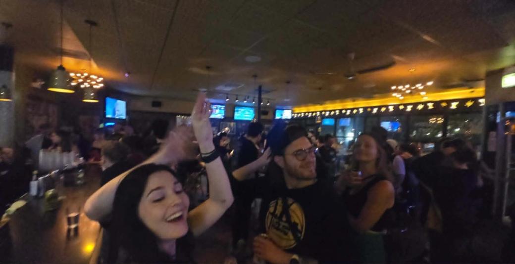 A crowd of people smiling in a dimly lit bar, with the bar counter on the left and TVs and yellow lights in the background. 