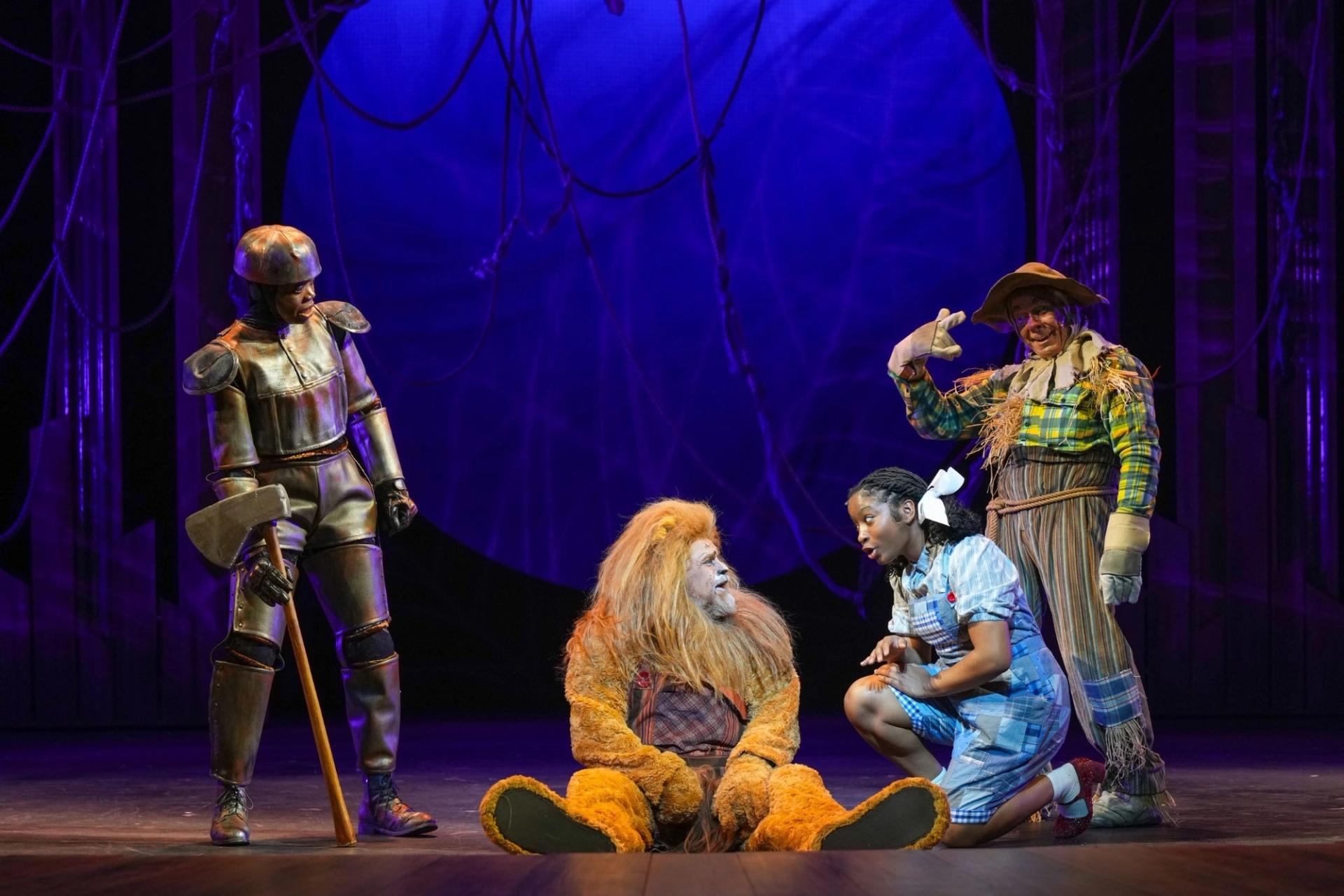 Four people on stage. On the left there's a man dressed in metal, next to him is a man dressed as the cowardly lion, next to him is a woman dressed as Dorothy, behind her is a man dressed as a scarecrow