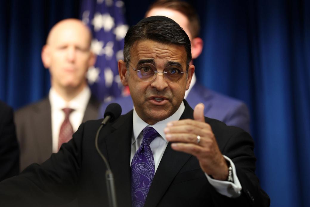 A close-up of man in a suit speaking in front of a microphone, with two men behind him.
