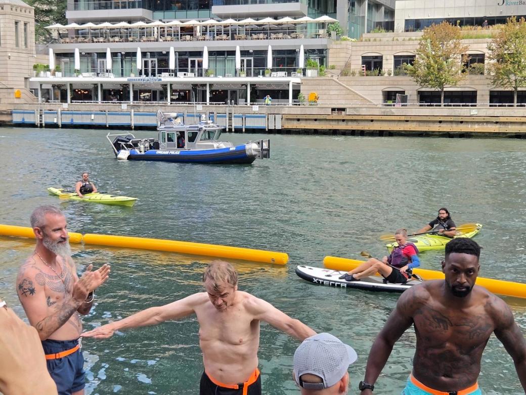 Three men with their shirts off by the river facing the camera. The man on the left has a grey beard and tattoos, the man in the middle is t posing and looking down, the man on the right has tattoos and is looking at the camera. In the background there are canoes and a boat.