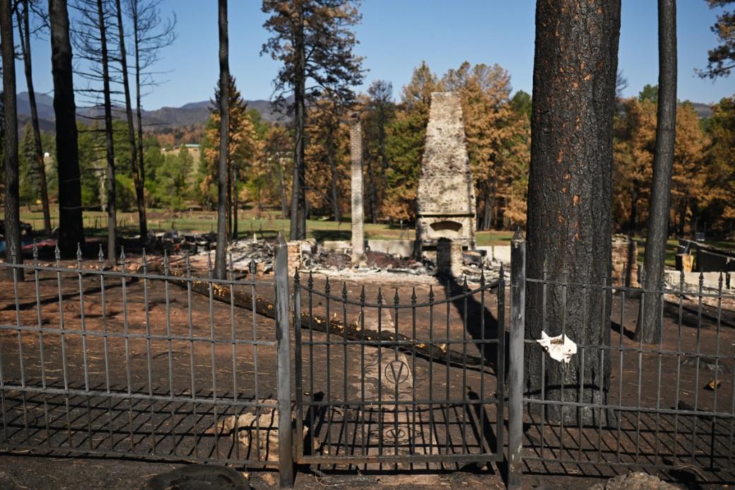 A stone fireplace and chimney rise from the burned remains of a home, behind an iron fence. 
