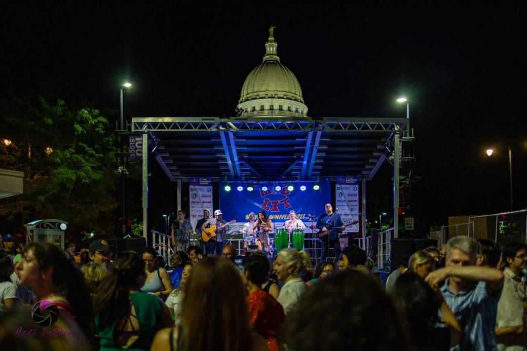 A concert stage is lit up in blue lights for a night concert in front of the Wisconsin State Capitol building.