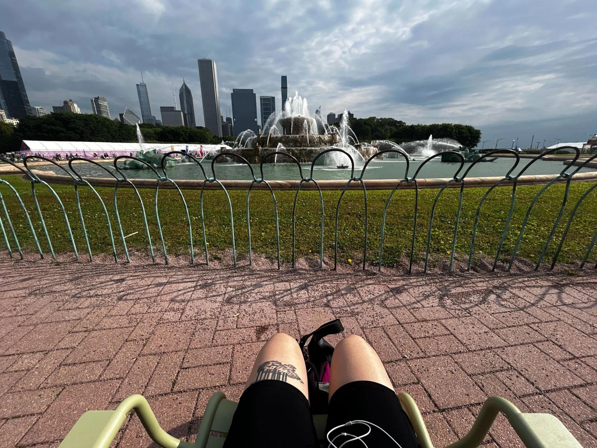 A point of view shot of someone sitting in a chair facing a fountain. There is a green fence preventing people from going on the grass