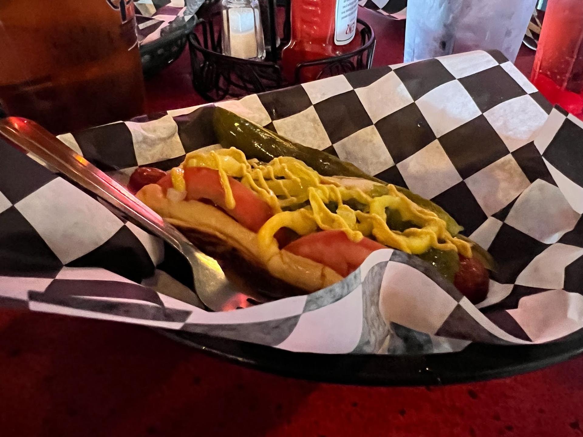 A hot dog with tomato, pickle, mustard, onions served on a checkered paper