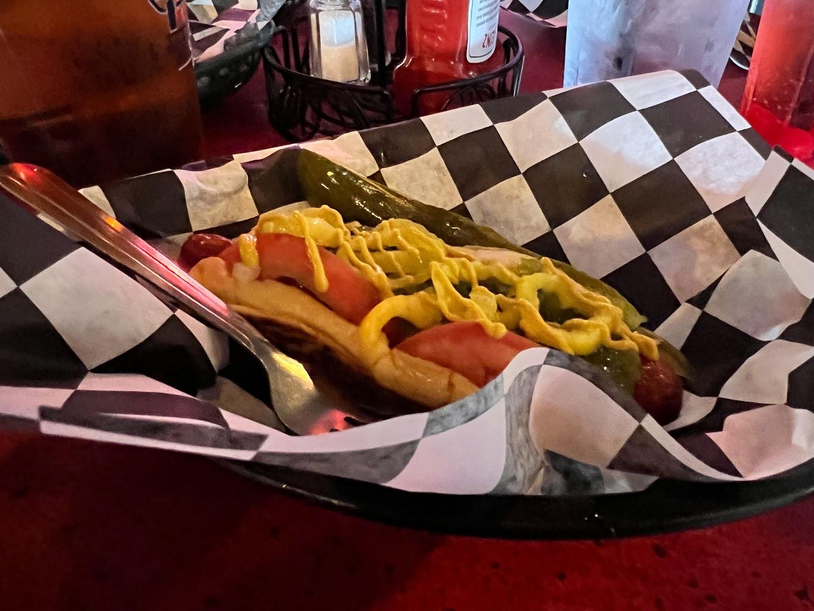A hot dog with tomato, pickle, mustard, onions served on a checkered paper