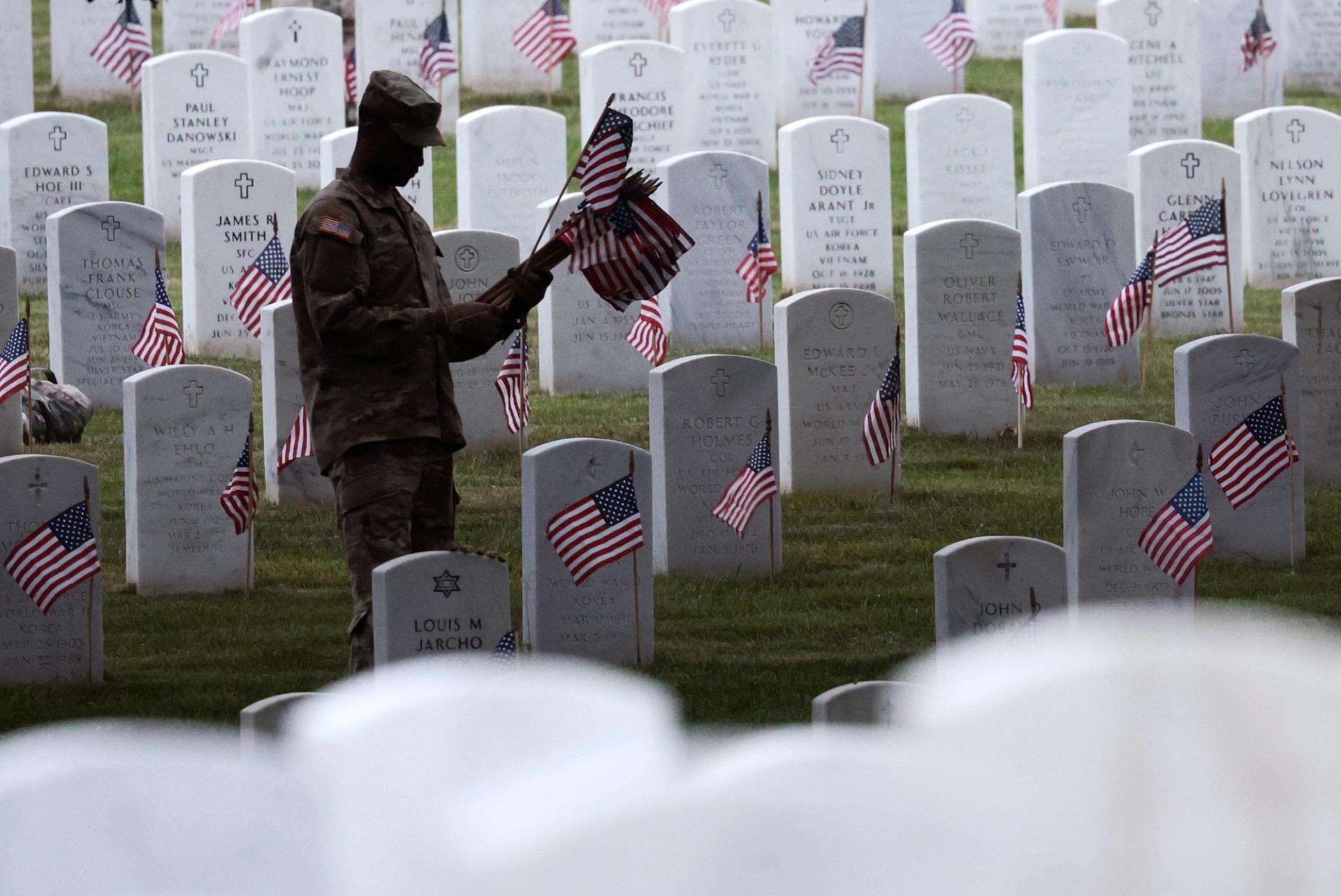 A soldier puts small American flags on rows of gravestones.