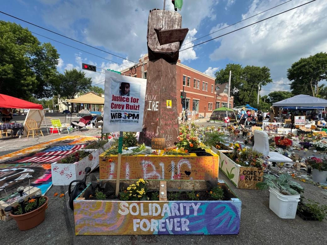 A makeshift memorial with flowers and candles and signs. One sign reads "Solidarity Forever."