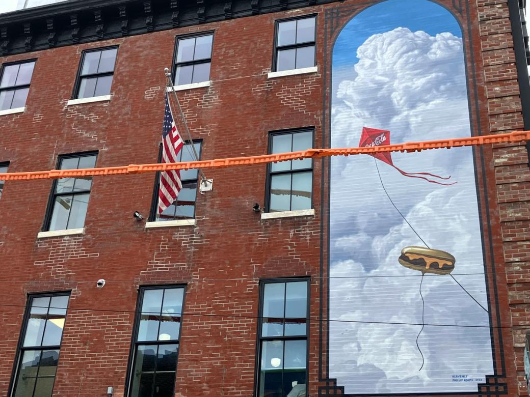  A brick wall of a building with multiple windows, an American flag, and a mural. The mural on the right-hand side is painted vertically across several floors, depicting a cheesesteak balloon and a Coca Cola-branded red kite floating among the clouds in the sky. 