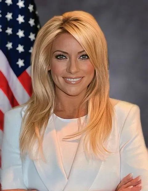 A pale woman with long blonde hair wearing a white suit smiles into the camera with her arms crossed over her chest. An American flag is visible in the background.