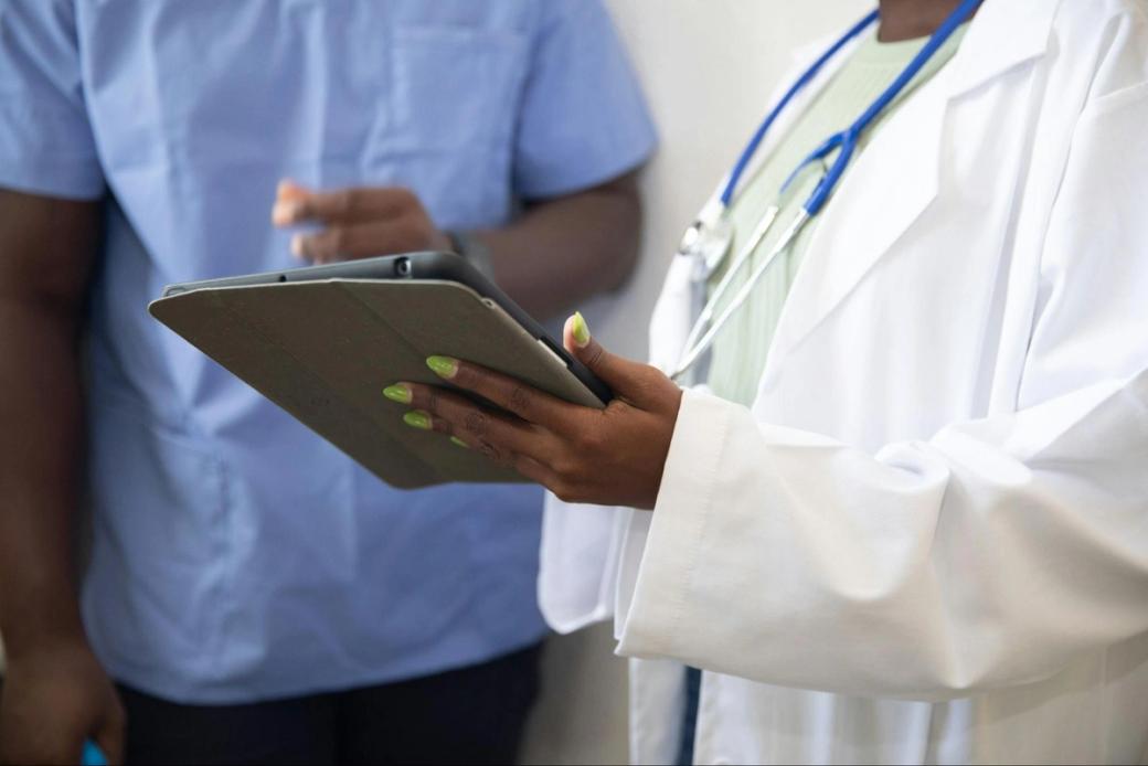 person in white coat, stethoscope, and with green nails holds a tablet. Next to a person in blue scrubs.