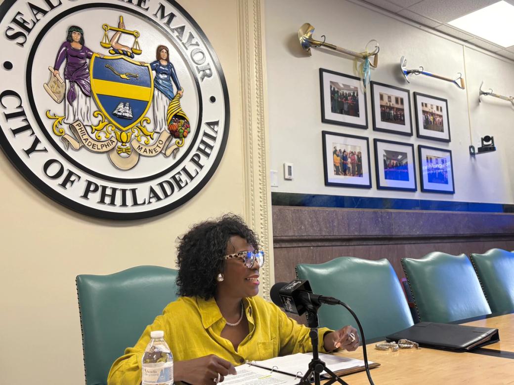 Mayor Cherelle Parker sits at a table in front of a mic. She is wearing glasses and a yellow blouse.