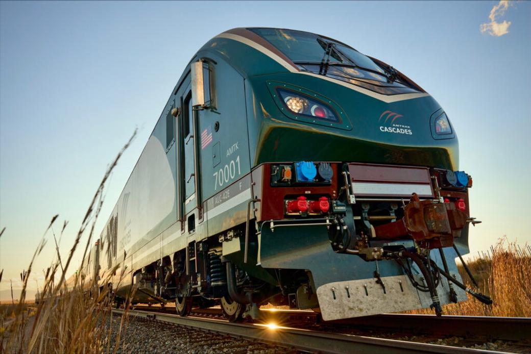 The new Amtrak Airos train is a deep green color with the “Amtrak Cascades” logo on the front and sides of the cars. The train glides past a wheat field during golden hour.