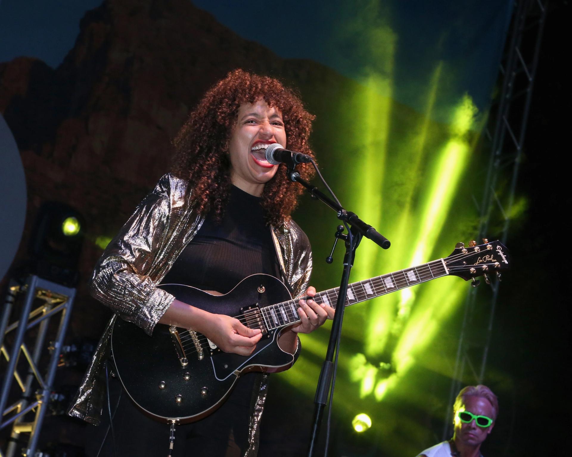 Jackie Venson playing a black guitar and singing into a microphone.