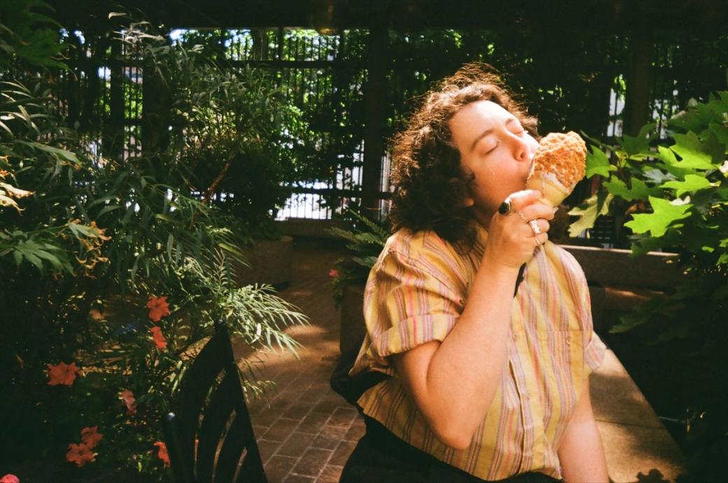 A person with brown curly hair licks a soft serve cone with her eyes closed while standing inside a lush green park in Pioneer Square