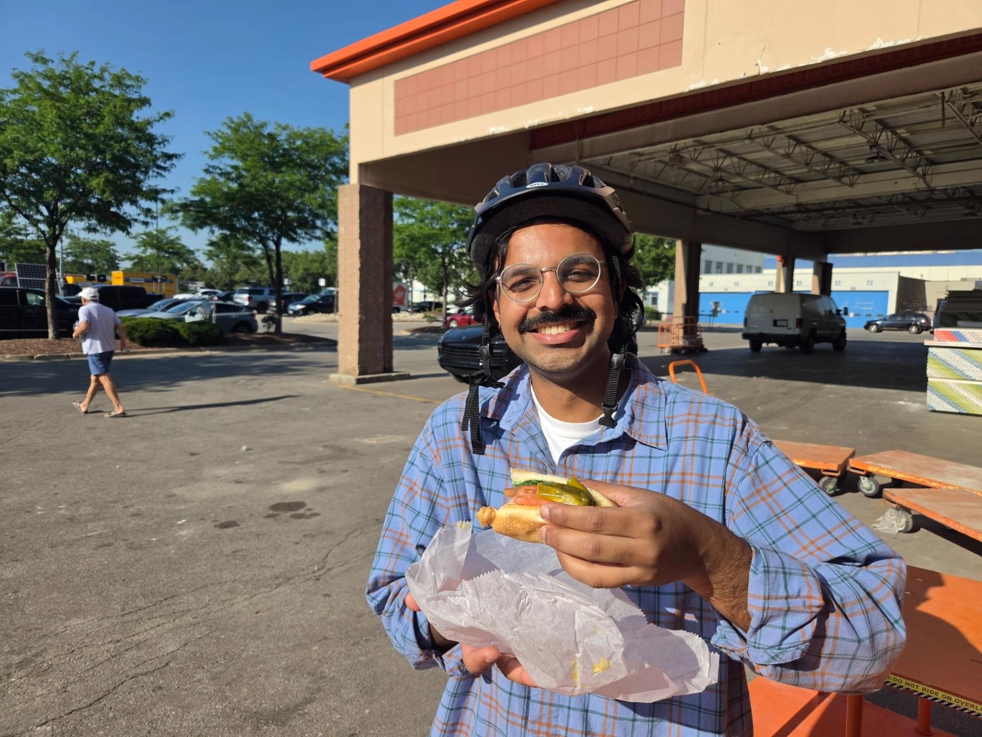 A man with a mustache wearing a helmet and glasses smiling at the camera and holding a hot dog