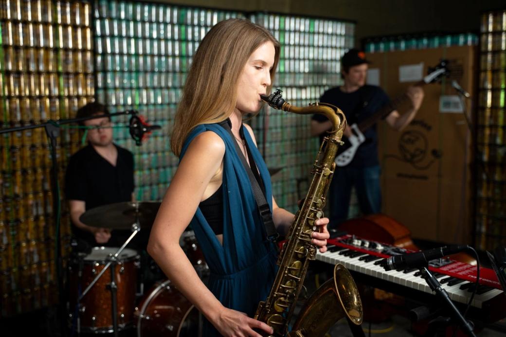 A woman plays saxophone with a drummer and bassist in the background. The band is set in front of a wall made of thousands of beer cans.
