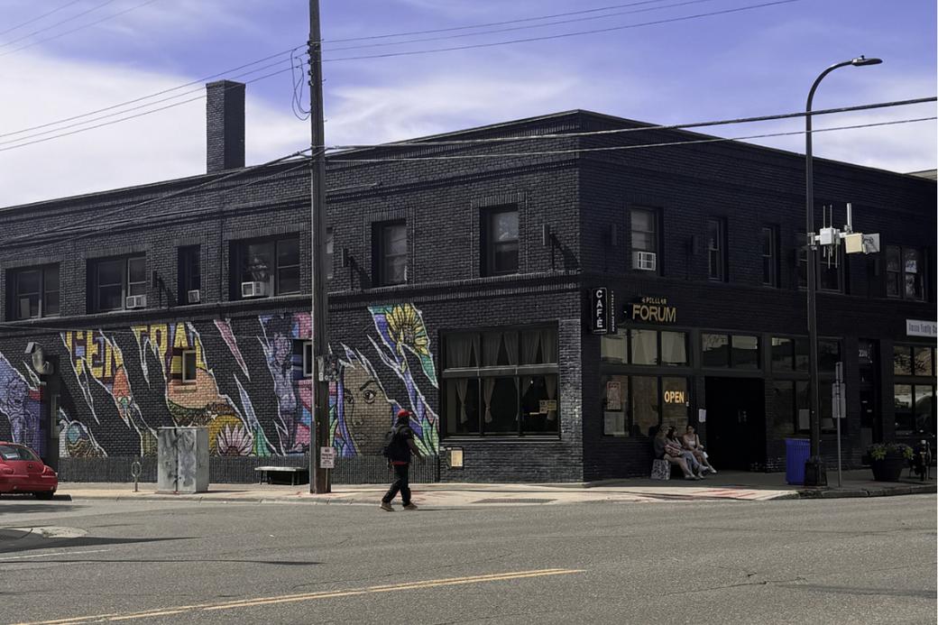 Exterior of black brick building with colorful mural on side, a pedestrian is crossing the street in front of the building