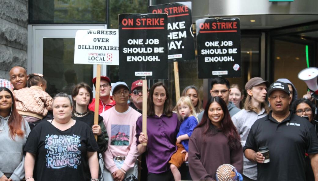 Local 8 Hotel workers stand alongside Mayor Wilson holding her daughter on her hip and Executive Zahilay holding his daughter as well. The Mayor holds a sign reading “On Strike. One job should be enough.” Two other people in the crowd hold the same sign and another holds a sign reading “Workers over billionaires.”