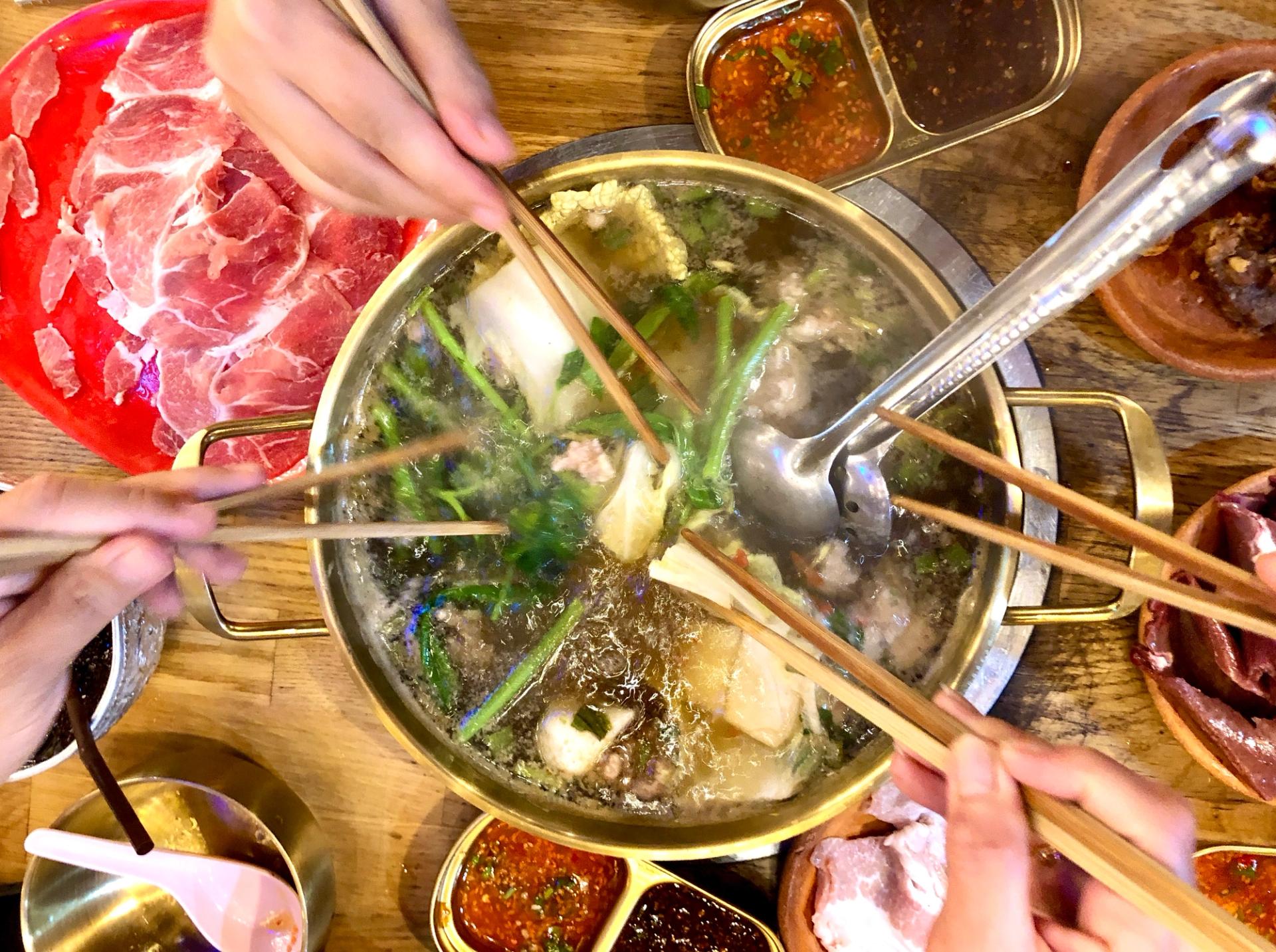 A hot pot with people's hands reaching over it and holding chopsticks.