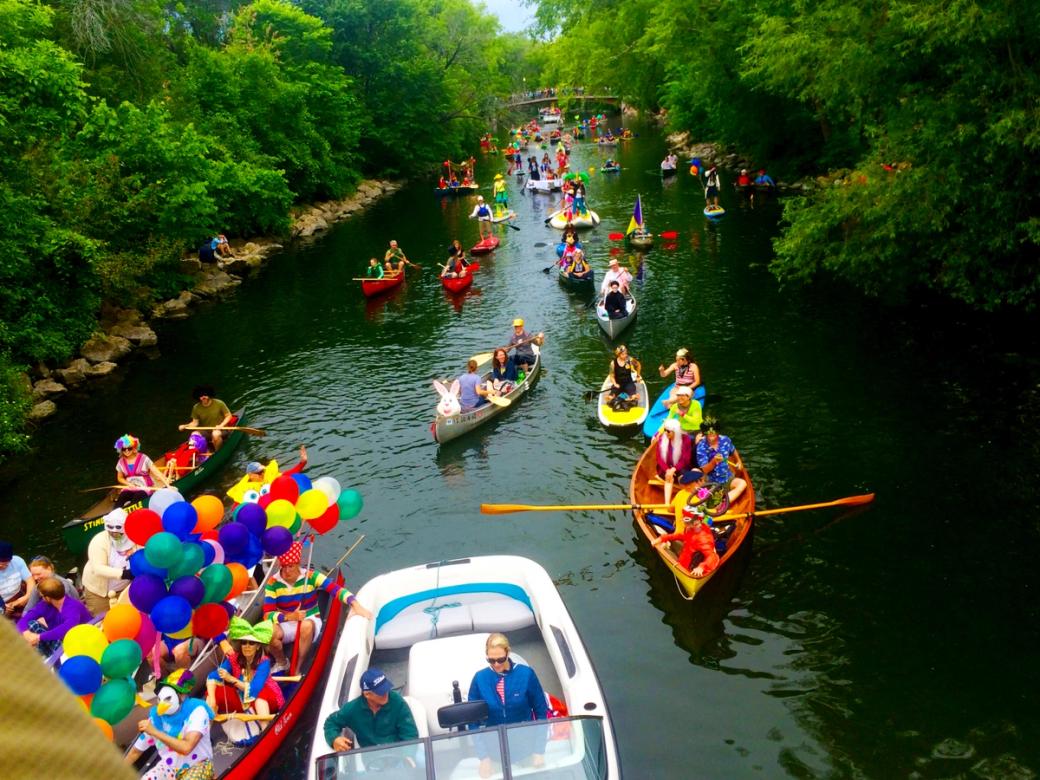 People in boats paddle and float down the Yahara River. One boat has balloons of different colors.