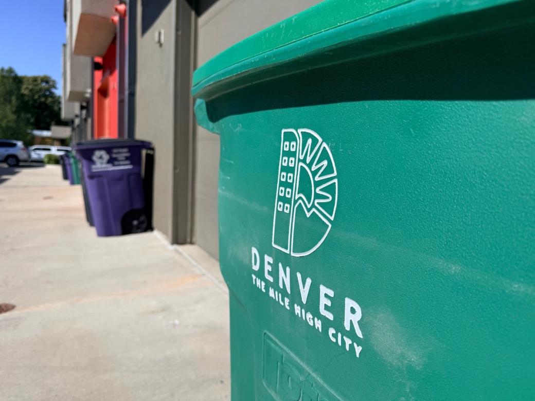 A green Denver compost bin in the foreground with a row of houses lining the alley in the background