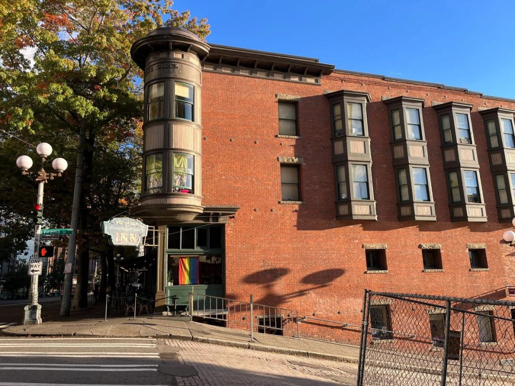 Photo of the Virginia Inn from the side facing Virginia Street on a sunny fall day in Seattle. The exterior of the building is orange brick with five picture window columns extending from the brick. The Virginia Inn sign is blue with white lettering and a rainbow flag is visible in the entryway window.