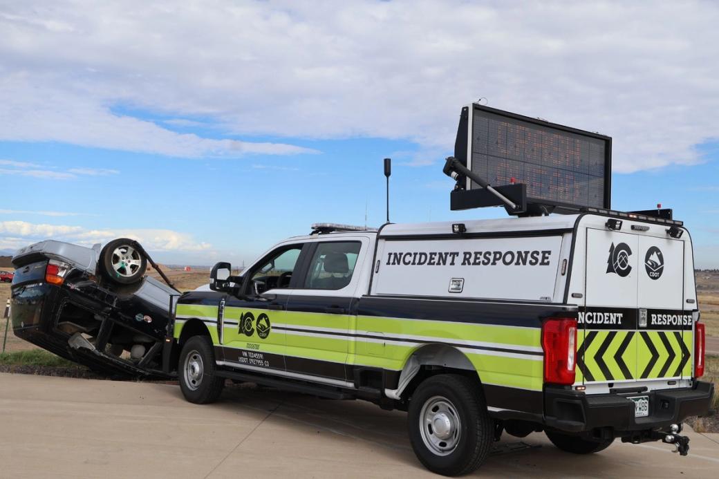 A neon green truck that says “incident response” on the side has a light up sign on top reading “crash ahead” as it is parked behind a flipped car.