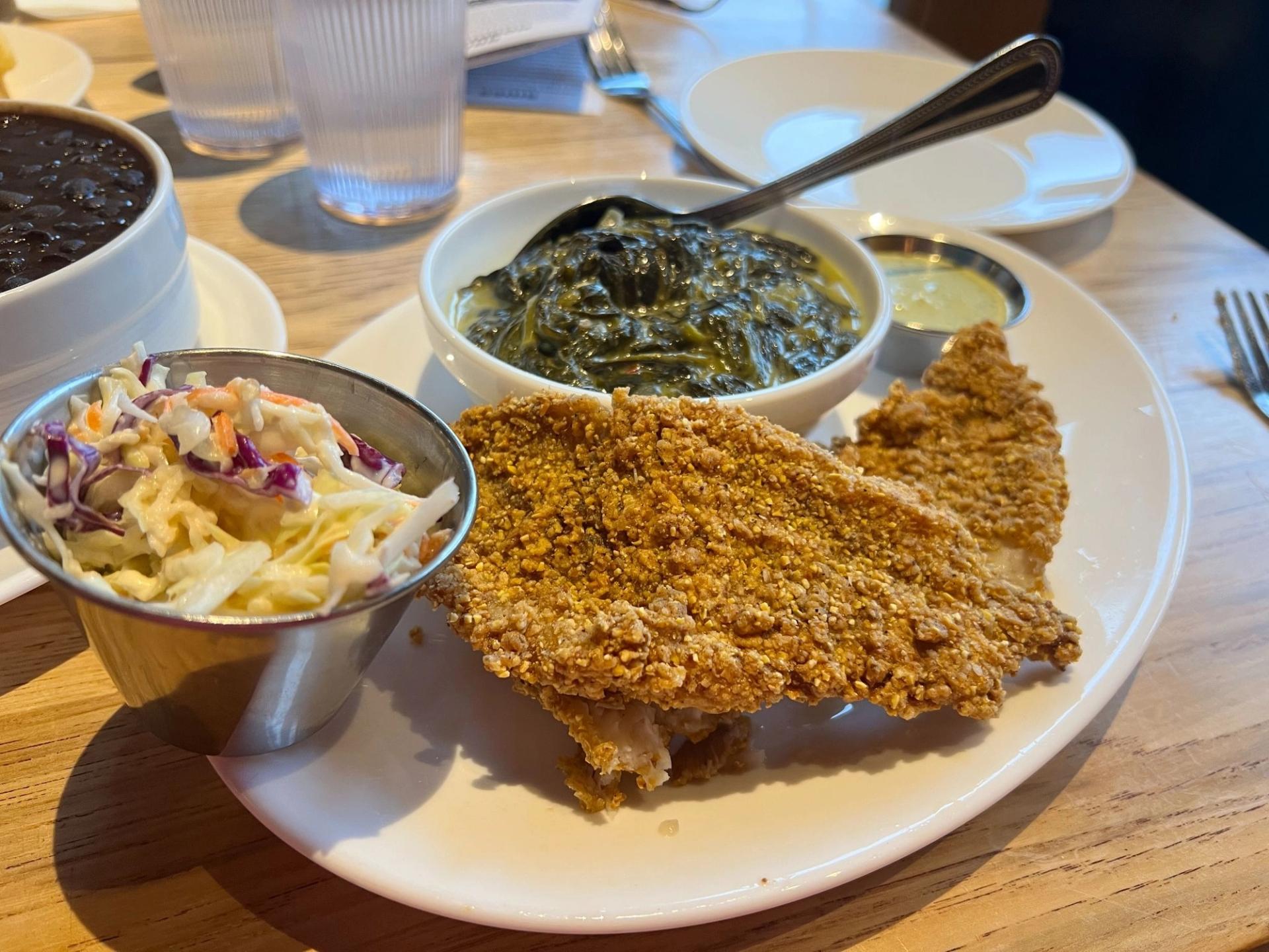 A plate with a deep fried catfish on it. There's a small bowl of creamed spinach, a small sauce container, and also a small bowl of coleslaw
