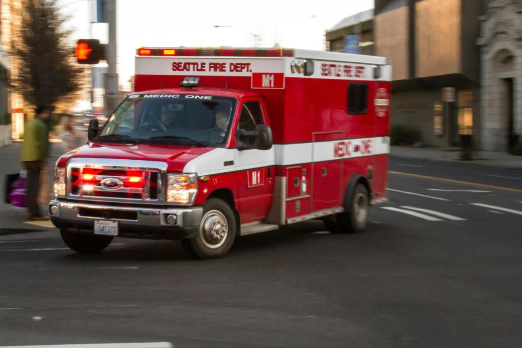 Red and white Seattle Fire Department truck turns a corner with its lights on in downtown Seattle. The background is blurred around it.