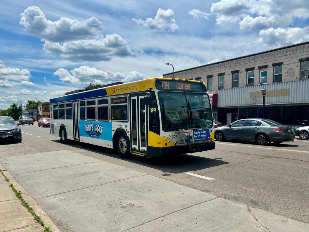 A blue and yellow bus on a street with other cars. A grey building and sidewalk are also visible.
